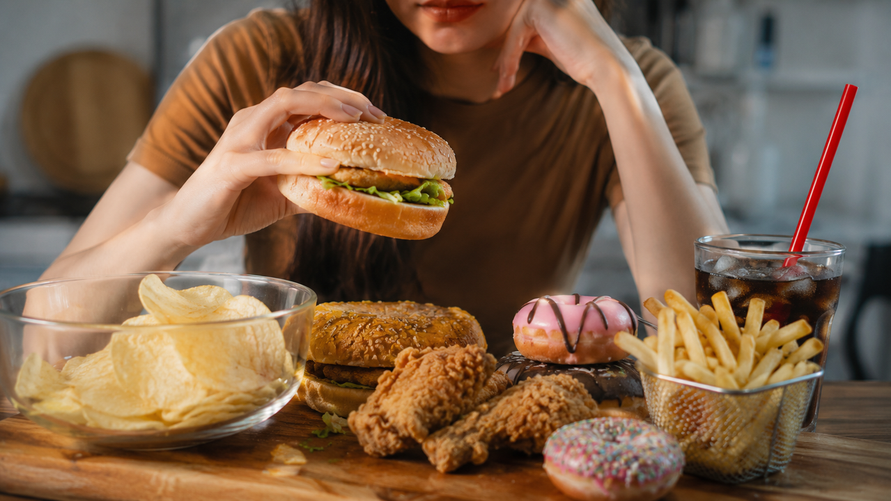 El Alimento Cotidiano Que Podría Estar Inflamando Tu Cuerpo Mujer sosteniendo una hamburguesa rodeada de alimentos ultraprocesados como papas fritas, refrescos, donas y snacks, representando hábitos alimenticios asociados con inflamación, cansancio y falta de energía.