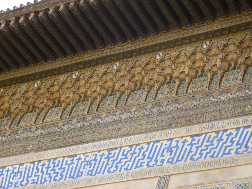 Carved underside of a roof.  There is white and blue tiles at the bottom.  Above them some very intricate stone work, with a trace of blue paint left. Above that is stone work in a larger pattern, all brown soft stone (probably sedimentary), then a green and yellow rail, followed by a dark wooden roof