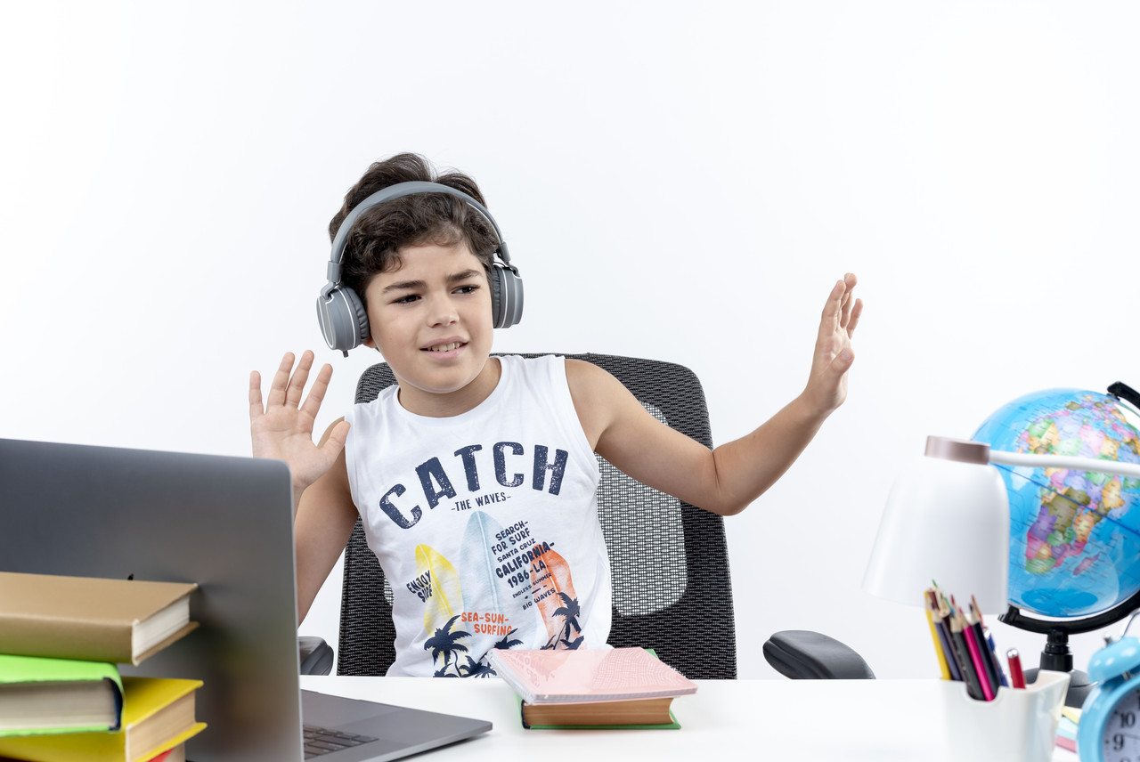 pleased little schoolboy wearing headphones sitting desk with school tools listen music isolated whi