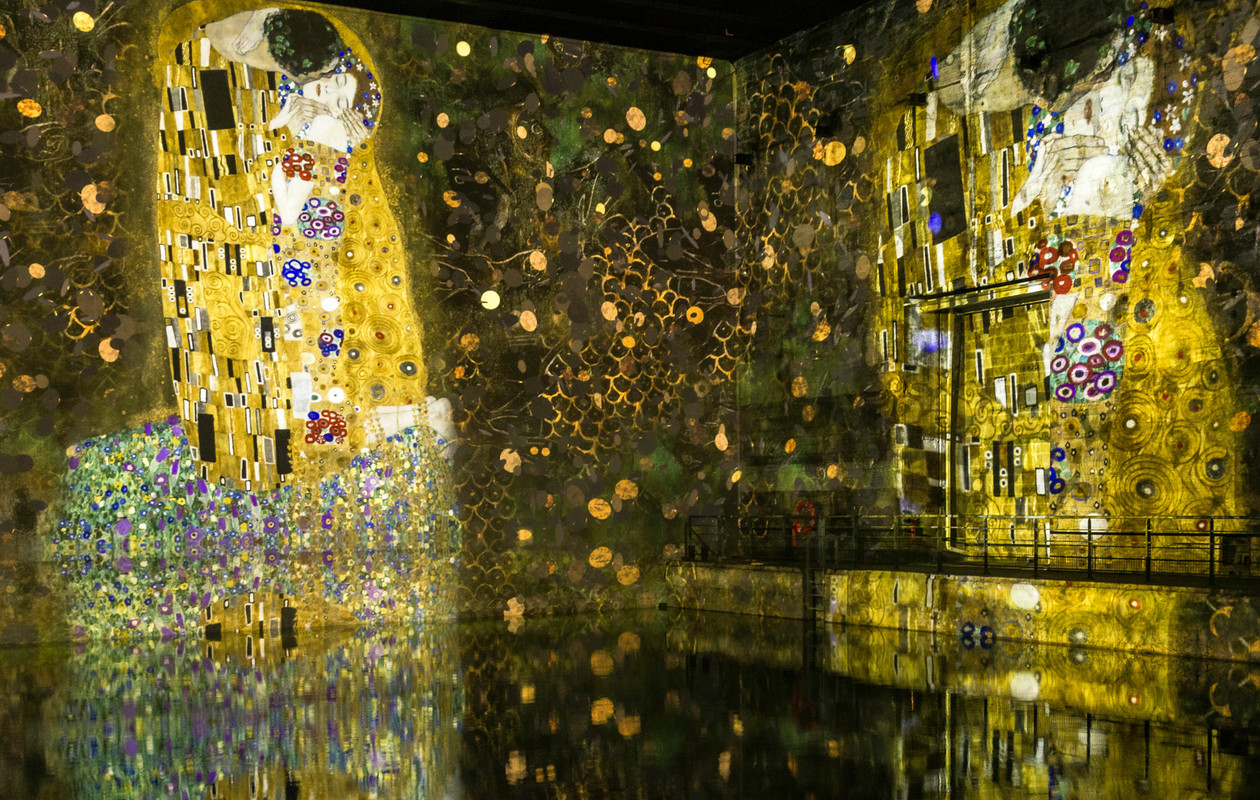 Immersive digital exhibition featuring Gustav Klimt's 'The Kiss' projected onto walls with golden hues and reflections in water.