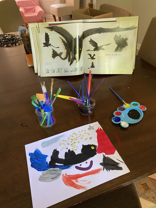 a desk with a watercolor painting of Toothless from How to Train Your Dragon. Below toothless is a purple happy face. It is clearly painted by someone very young. An art book reference is also pictured.