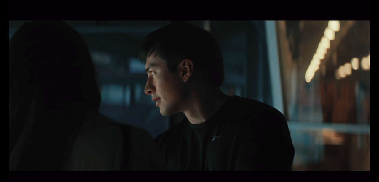the back of yuna's head fills the left side of the frame. in the center is shane, facing towards yuna and looking to the right so he's in profile, face lit by the lights from below. he's wearing a long-sleeved black shirt.