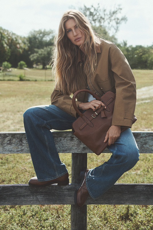 Gisele Bündchen, con i capelli sciolti, indossa un completo di jeans a gamba larga e giacca di tela color cammello, mentre posa seduta su una staccionata