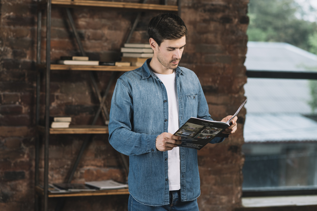handsome young man reading magazine standing front book shelf