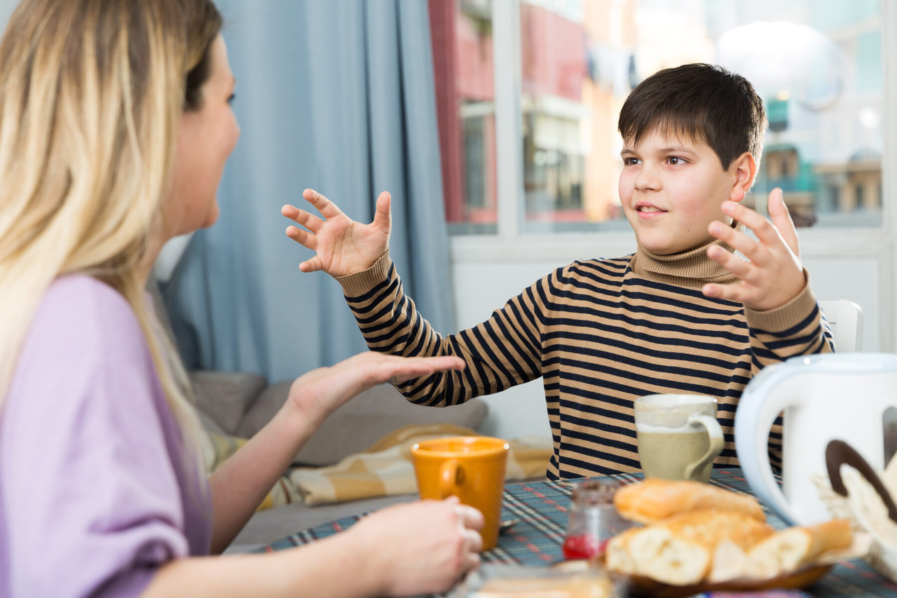 A boy gestures animatedly while telling his mother about something at the breakfast table.