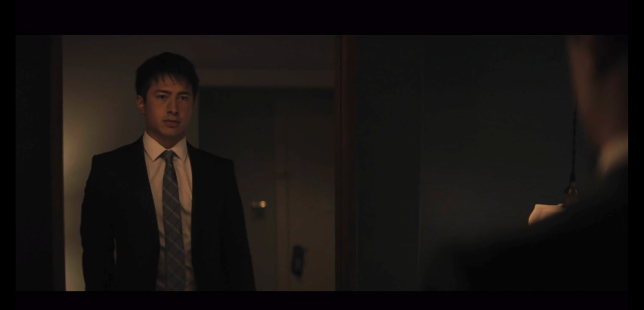 shane looking at himself dressed in a suit in the mirror. the shot is framed so we're looking over his shoulder into the mirror. he's in a black suit jacket, white shirt, and silvery-blue  tie with criss-crossing stripes.