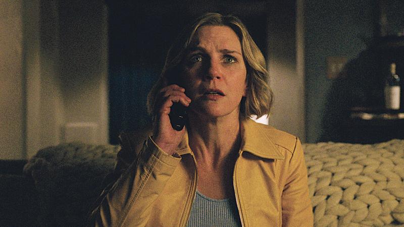 a still frame from episode 1 of pluribus. carol is sitting on the couch in her home and talking to the hivemind by speaking into the telephone and hearing its response through her TV