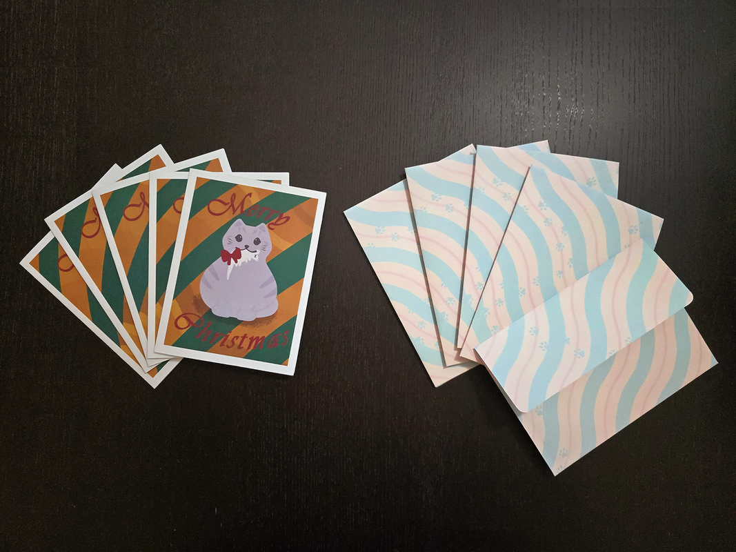 A small stack of Christmas cards, a small stack of envelopes with the back facing the camera, and a single envelope with the flap facing the camera. The cards have a round grey cat holding a dead mouse with a red bow on the mouse in the cat&#39;s mouth. The cat is standing in front of a gold and dark green striped background. The words &quot;Merry Christmas&quot; are printed on the card in maroon cursive. There is a white border around the edge of the card. The envelopes have parallel squiggly stripes diagonally covering the envelopes. The stripes are a repeating pattern of thick light green, medium thickness light yellow, thin light red, and medium thickness light yellow again. There is a path of paw-prints walking across the envelope in the same light green as the diagonal stripes. The envelope with the flap facing the camera is printed with the same pattern and reveals the flap to be a flat-edge flap with rounded corners.