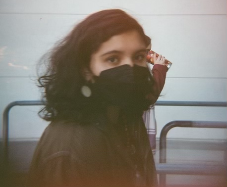 a photo of a light-skinned but brown person with medium-length brown hair walking looking to the side at the camera. they have a black facemask, circular dangly earrings, and a brown button-up  on, but the photo seems to have a filter on it that makes them look warmer and the background look cooler.