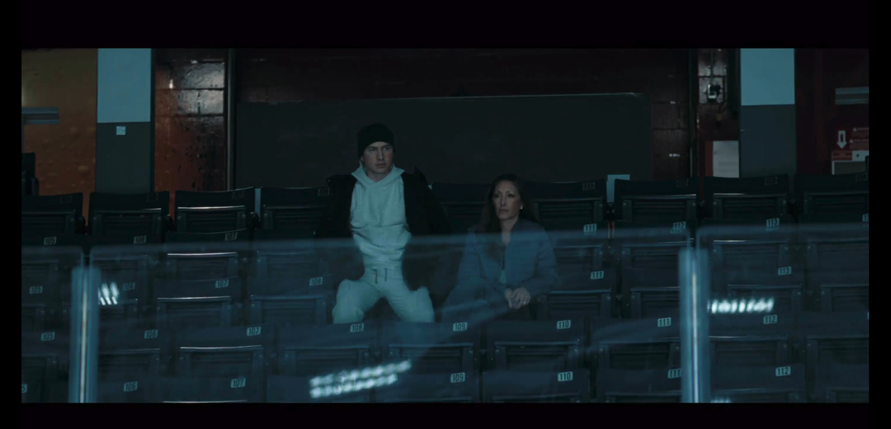 shane and his mom sit centered in the arena stands. shane is perched on top of an arena chair with the seat flipped up. his mom is sitting properly in the seat. shane is wearing a dark green beanie, white sweatshirt, black button-up coat, and white sweat pants. shane's mom, yuna, is wearing a blue-gray coat.