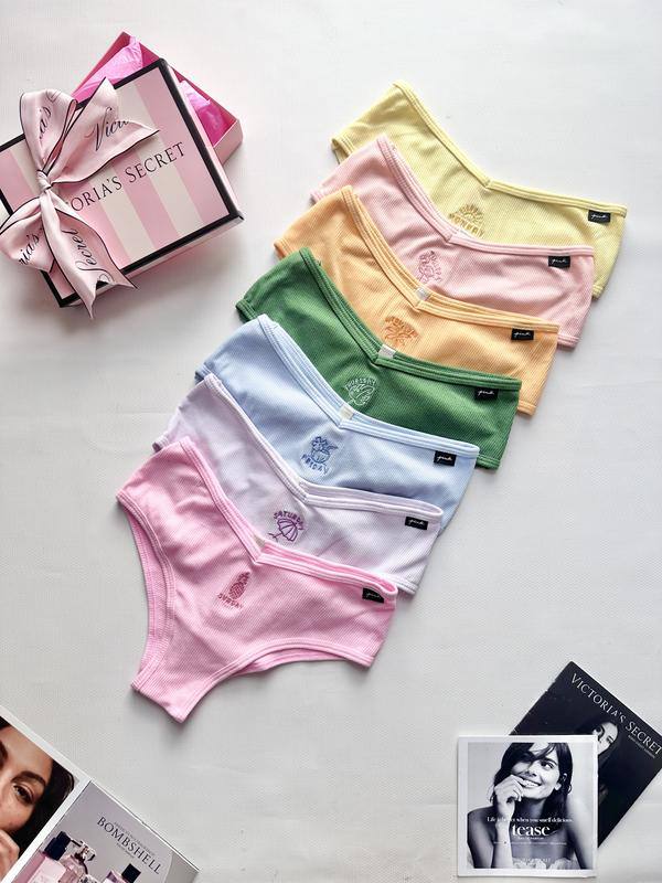 Разноцветные женские трусики из рубчатого трикотажа (розовые, белые, голубые, зелёные, жёлтые и бежевые) разложены стопкой на светлом фоне рядом с подарочной коробкой Victoria’s Secret и журналами.