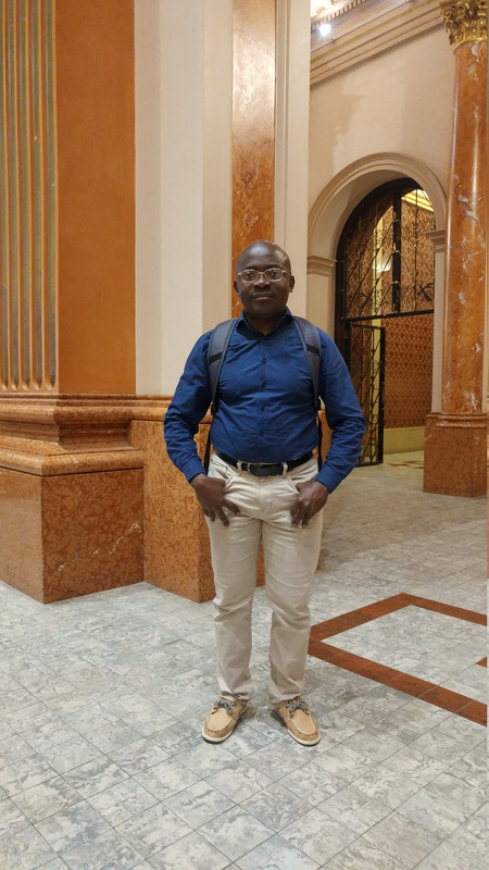 A full-length photo of Dr. Arnaud Tcheutou standing inside an ornate building featuring grand architecture. He is wearing glasses, a long-sleeved dark blue button-down shirt, beige trousers, and light brown shoes. He carries a dark backpack over his shoulders and stands facing the camera with a warm expression. Surrounding him are thick, warm-toned orange marble columns and an arched entryway leading into an adjacent room with decorative walls.