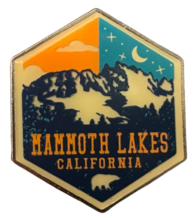 a hexagonal enamel pin for mammmoth lakes california with dark blue mountains in front and an orange daytime sky on the left side & a blue nighttime sky on the right