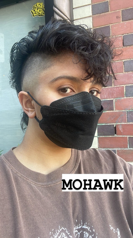light skinned brown person [me!] with a black mask on, a brown shirt, and a new mohawk haircut! zie are facing slightly to the right of the camera, but looking back at it