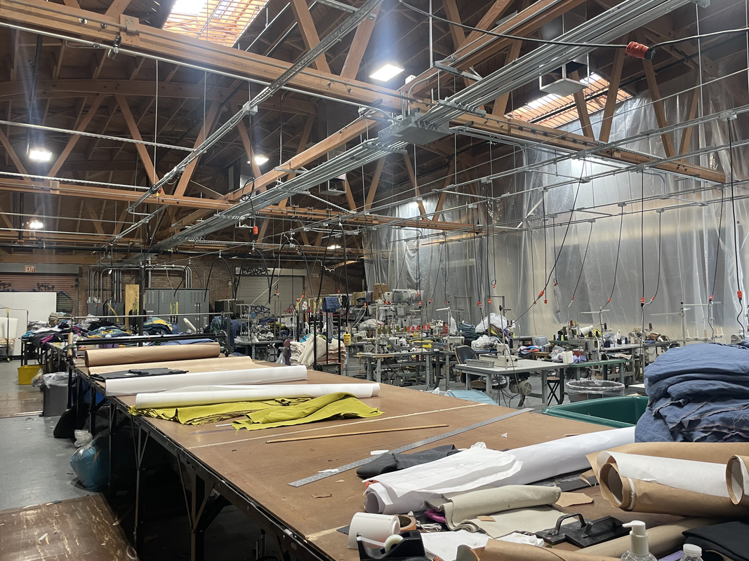 A photo of a large sew shop, with lots of tables and equipment. The entire place as wooden and has a lot of beams on the ceiling. There are a few skylights that let a lot of light in and stream down, making it look sort of holy.