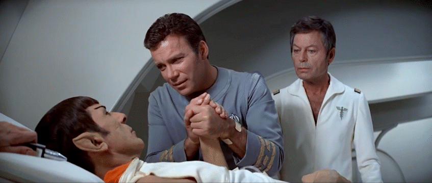 in this gif are Leonard Nimoy's Spock, William Shatner's Kirk, and DeForest Kelley's Dr. McCoy. Spock is lying in a hospital bed and holding Kirk's hand