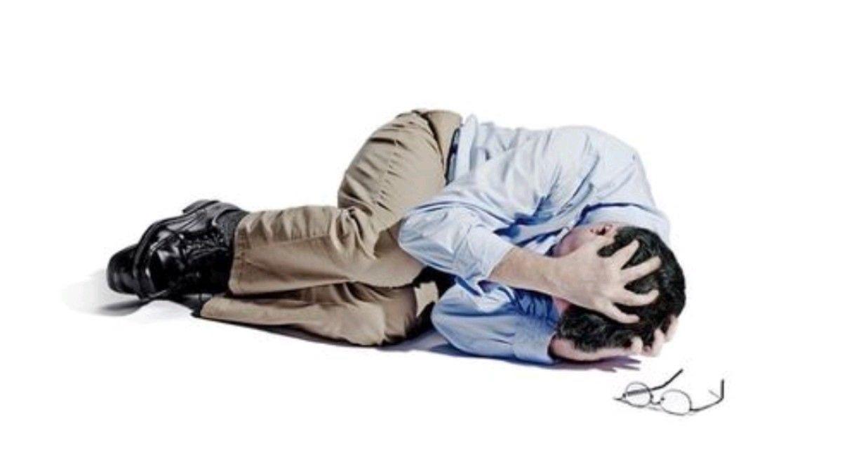 Stock photo of a person in business-casual clothes curled up in the fetal position on the floor of a white void gripping their head.