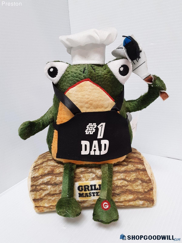 a product photo of a stuffed frog sitting on a log. the frog is wearing a chef hat and holding a spatula with a fly on it. the frog is also wearing an apron which reads, 'number one dad'. the log is labeled, 'grill master'