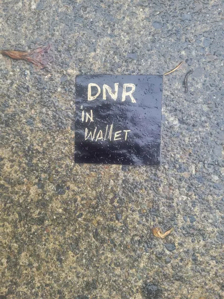 a photo of a black sqaure on concrete the sqaure has gold text on it that read DNR in wallet. Both the sqaure and the concrete are wet from rain and there is small debris that looks like plant leaves as well as dirt on the concrete.