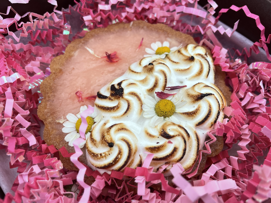 a photo of a light pink tart with darker pink flowers on it, and meringue covering one half of it. it rests on a lot of light pink, hot pink, and red crinkle paper.