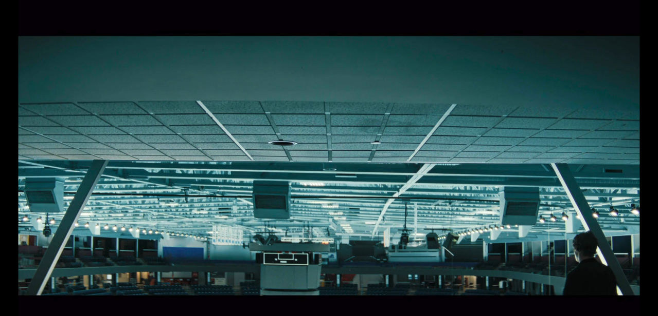 ilya is facing away from the camera and is in the bottom right corner of the composition. he's small in the composition. the rafters of a skating rink fills the rest of the image.
