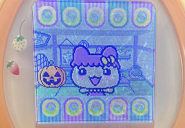 close up photograph of a tamagotchi plus color device. chamametchi is on the screen, close to the viewer, and posing in a slightly different cute pose than in the first image. a festive jack-o-lantern is in the room with her.