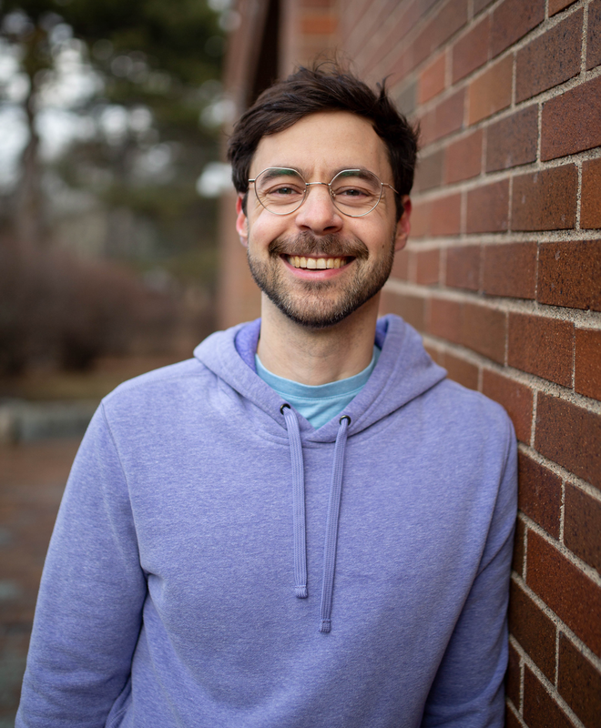 Ben standing next to a brick wall, smiling, wearing glasses and a purple hoodie