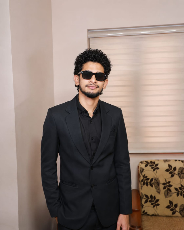 Muhammed Lisan A in a black suit and sunglasses, standing indoors