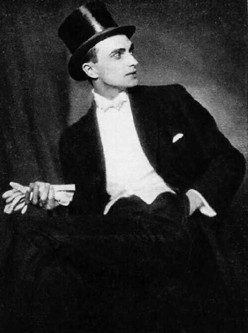3/4 length black and white portrait of German-British actor Conrad Veidt, circa 1920, in white tie formal wear and top hat, seated in front of a curtain and holding a pair of white gloves in his right hand, his left hand in his pocket, looking off camera in profile to the right