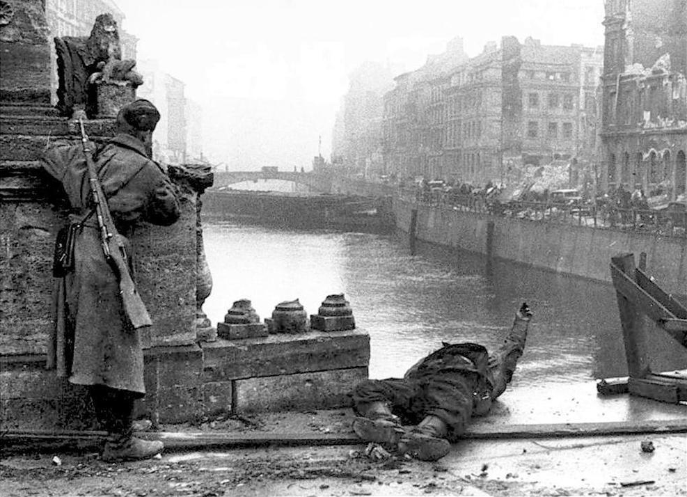Un ruso se asoma lentamente sobre un puente, mientras a sus pies se encuentra el cadáver de un combatiente alemán