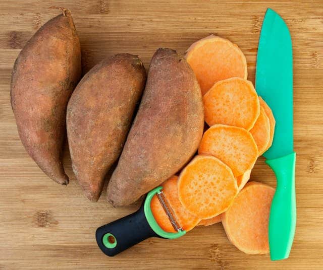 Image of whole and chopped sweet potatoes with knife and peeler placed on a cutting board